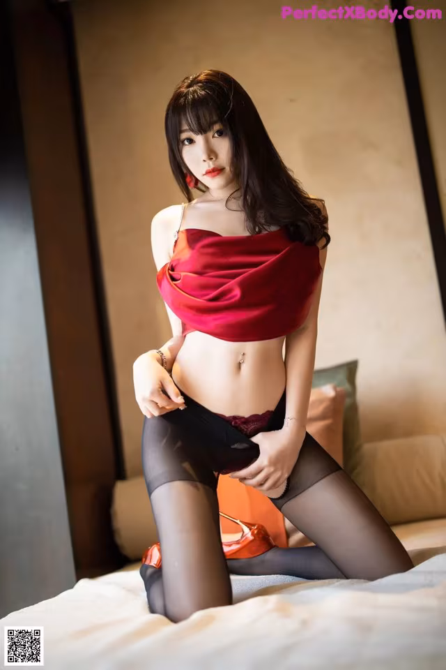 A woman in a red top and black stockings sitting on a bed.