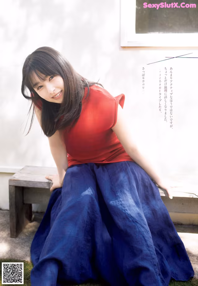 A woman sitting on a bench in a red shirt and blue skirt.